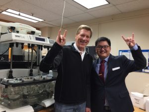 Dr. Williams and research trainee smiling in front of lab equipment and giving 'hook em horns' hand sign