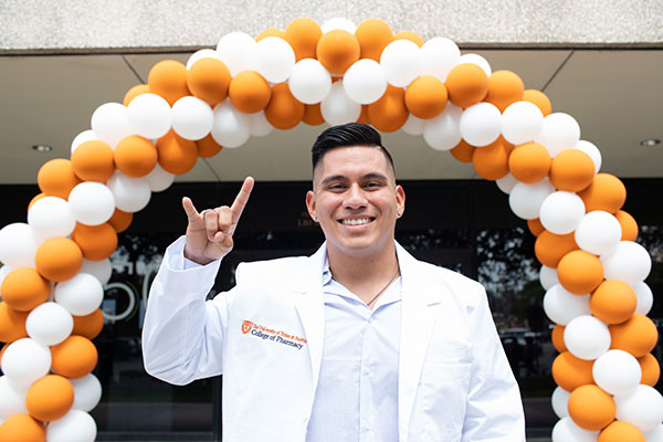 Student at white coat ceremony giving hook em horns hand sign and standing under ballon arch