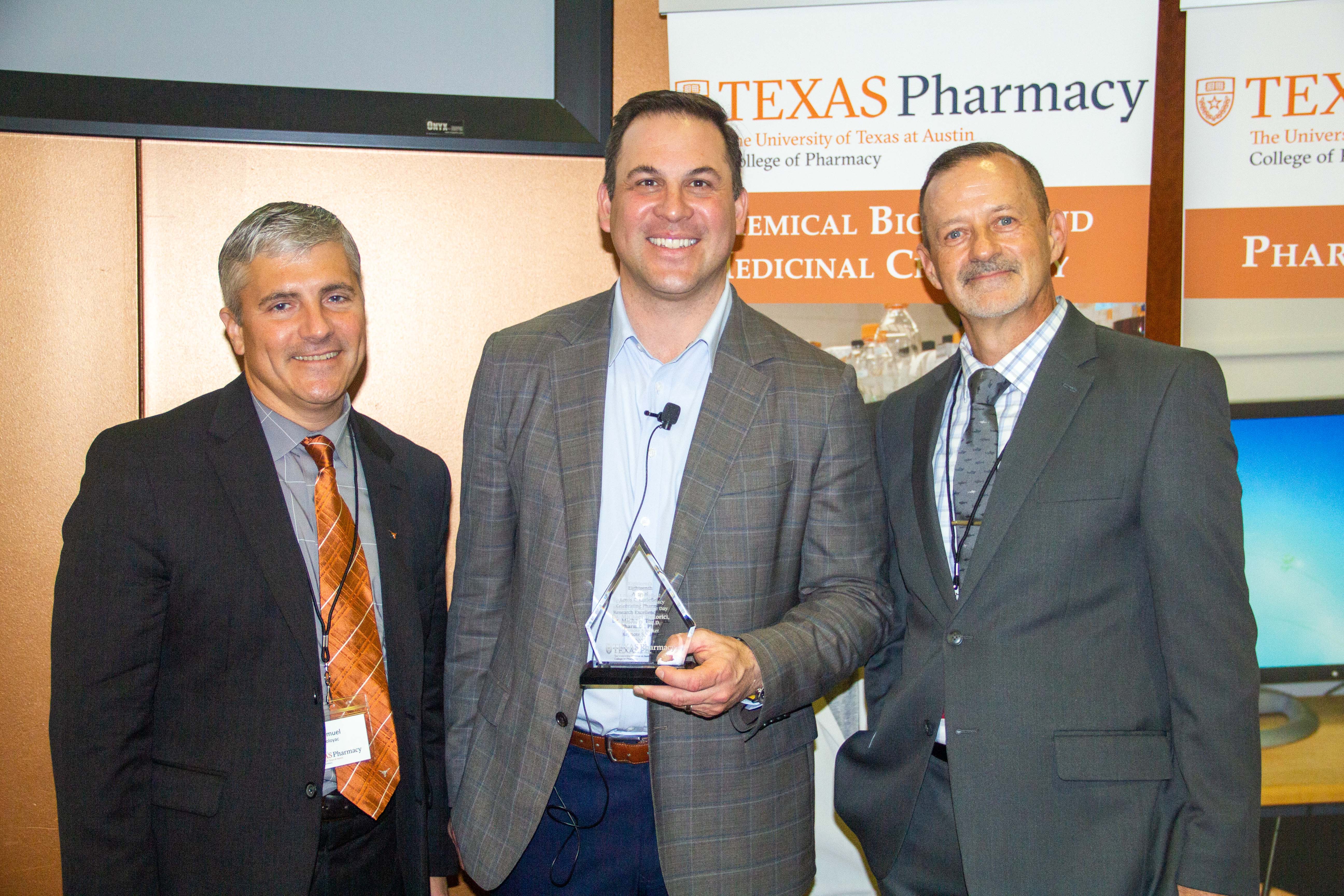 Three men wearing suits smiling for the camera. The man in the middle is holding a trophy award.