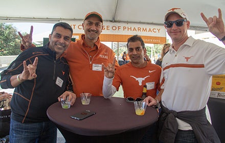 Four Pharmacy alumni smiling and giving hook em horns sign at cocktail table at Homecoming Tailgate event