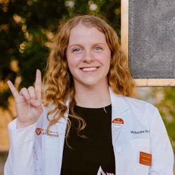 A woman smiling while wearing a white coat and giving the Hook 'em Horns hand gesture.