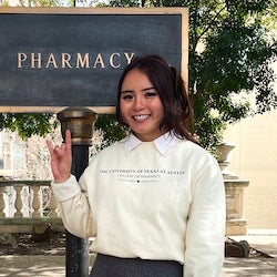 Jennifer Tran giving the Hook 'em Horns hand gesture in front of a sign that says Pharmacy.