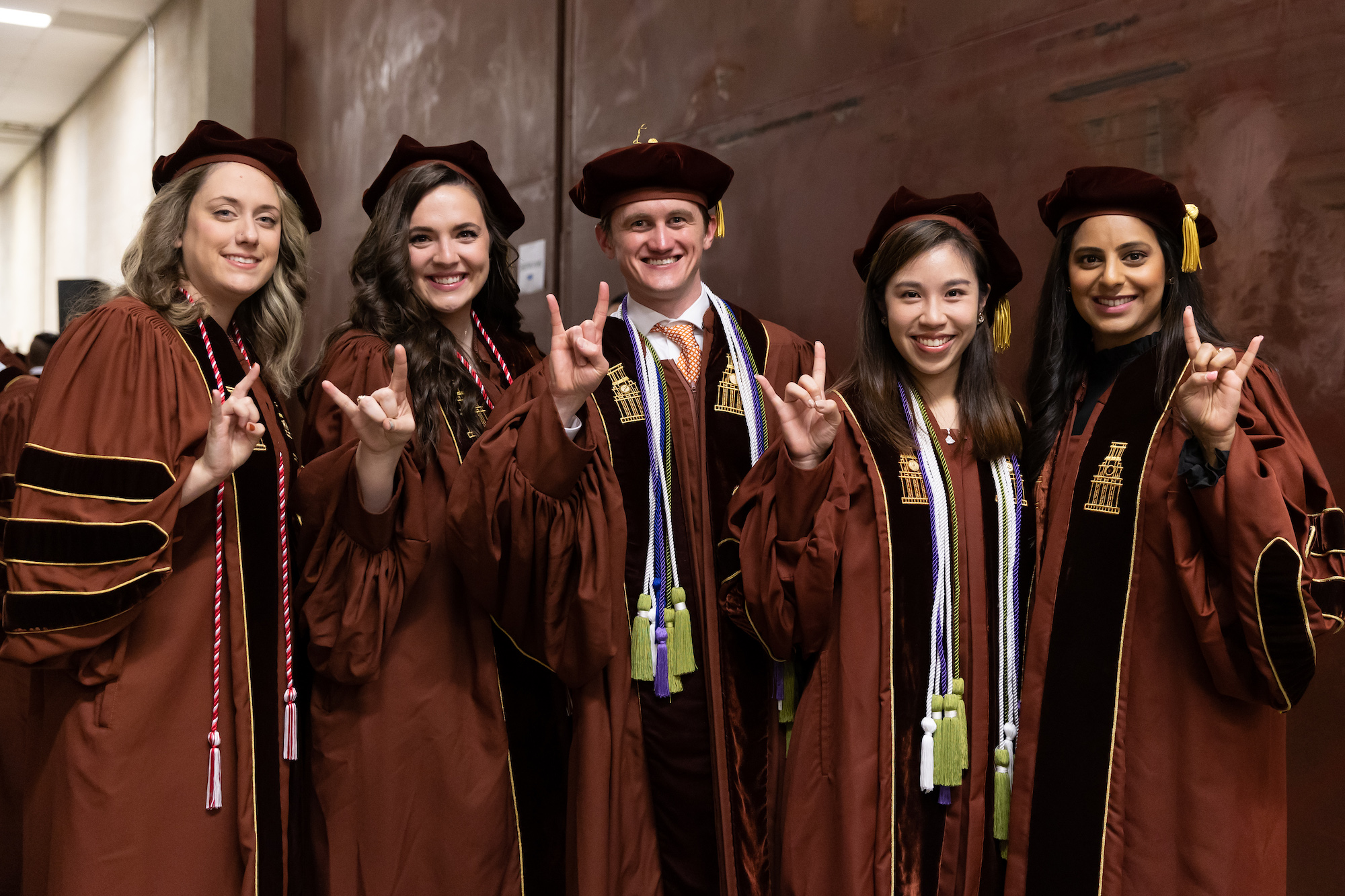Five students wearing graduating caps and gowns and giving the Hook 'em Horns hand gesture while smiling.