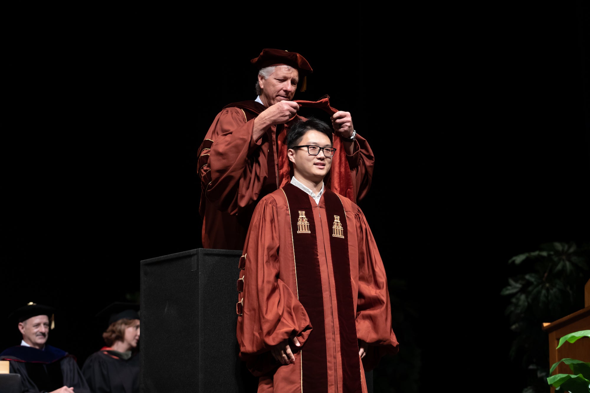 Two people in graduation caps and gowns, one is hooding the other.