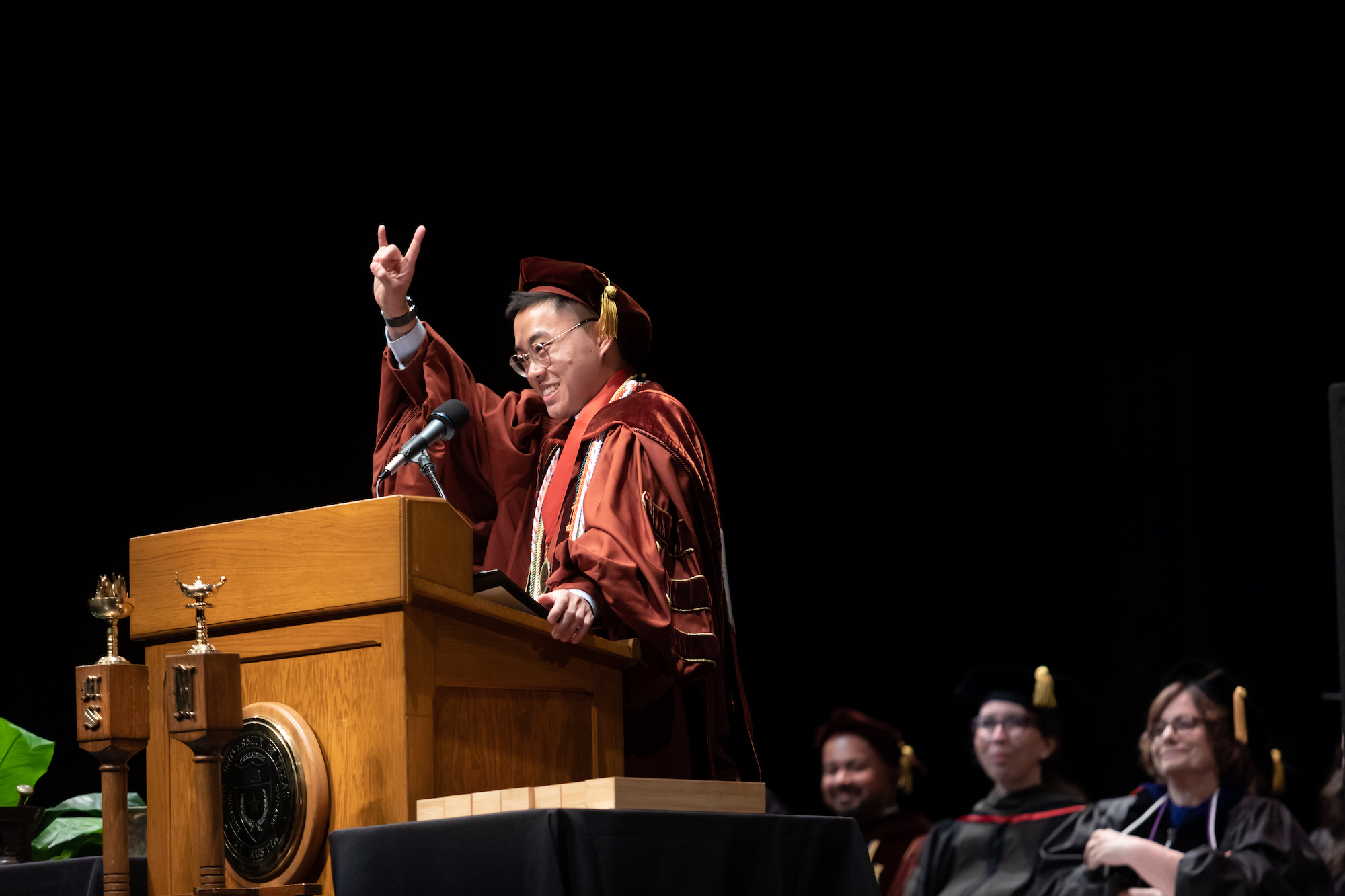 A man wearing a cap and gown at a podium and giving the Hook 'em Horns hand gesture.