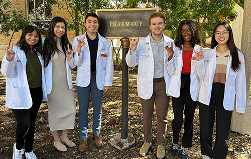 Six Pharm.D. students wearing white coats and giving the 'hook em horns' hand sign by the Pharmacy building sign