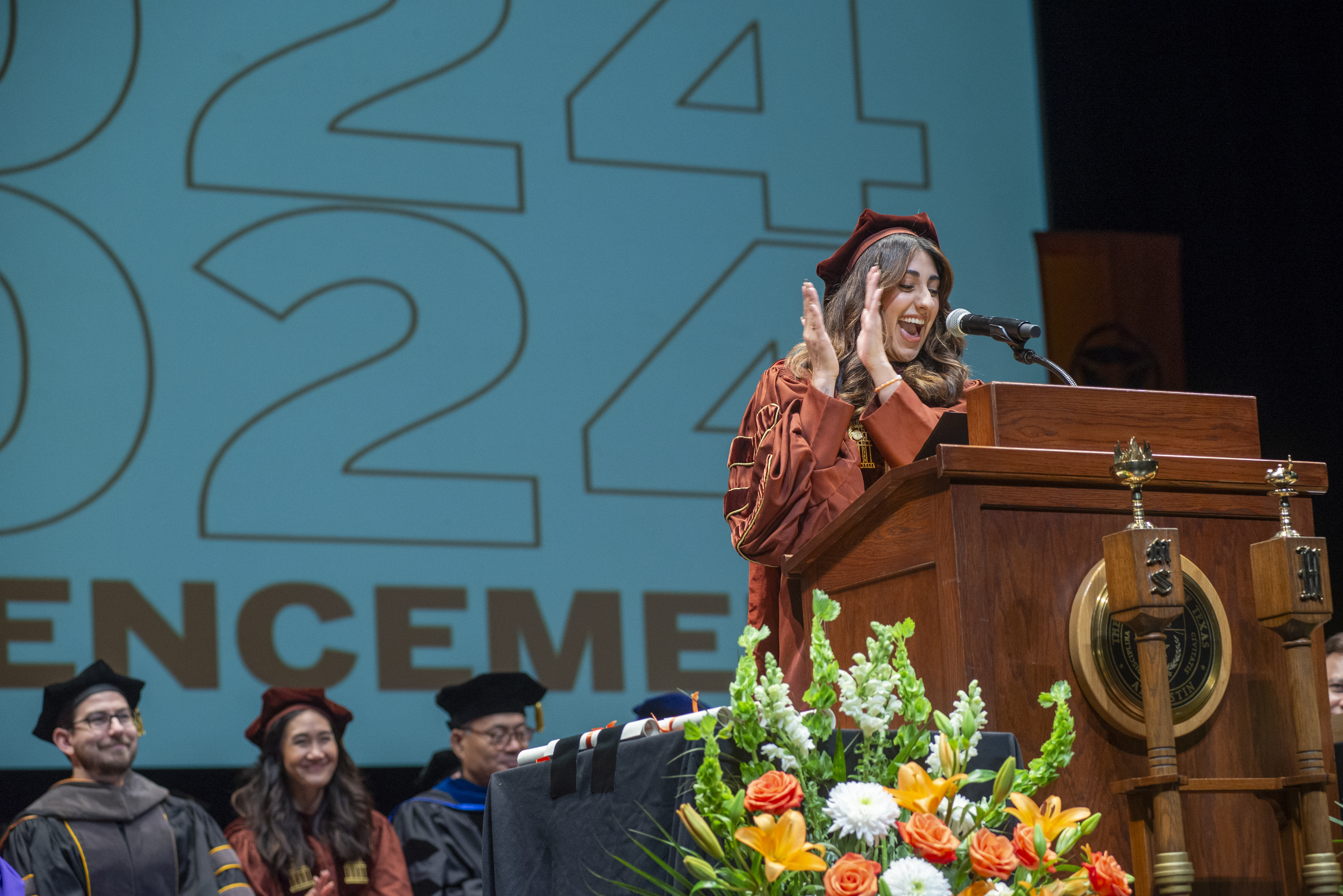 A woman wearing regalia and clapping while giving a speech in front of a podium.