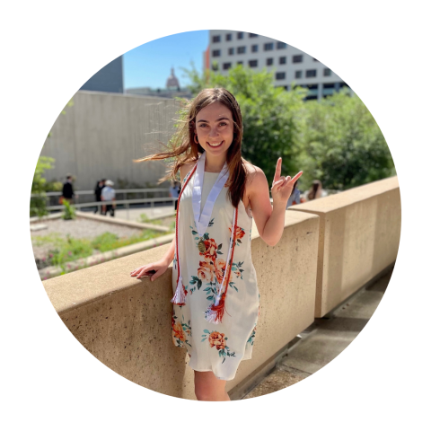 Erica wearing a floral dress and academic cords around her neck smiling to camera with a hook 'em (hand gesture) in the air.