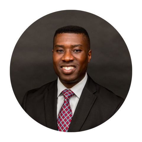 Emmanuel Enwere wearing a black suit jacket and red and blue plaid tie smiling to camera.