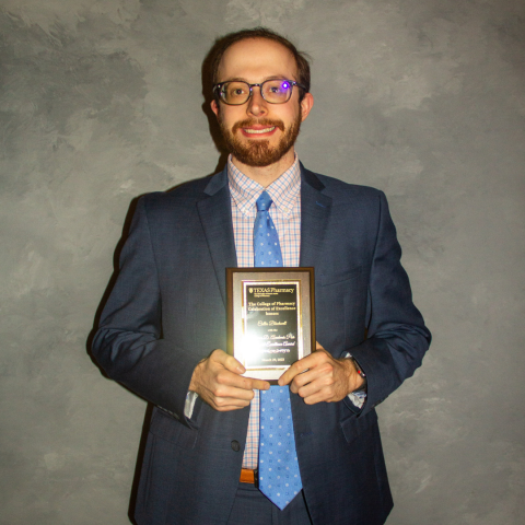 Collin Blackwell holding his award plaque. He is wearing round glasses, a navy suit and blue tie.