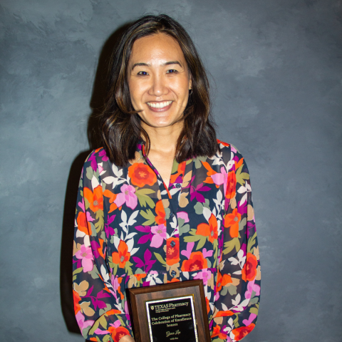 A photo of Dr. Grace Lee holding her award plaque. She has straight, shoulder-length brown hair and is wearing a multi-colored floral-patterned dress with long sleeves.