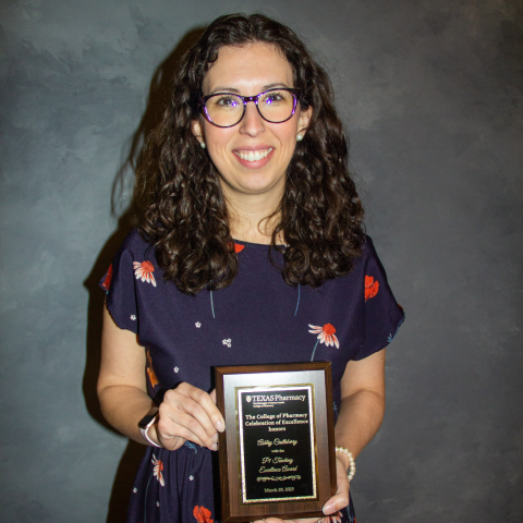 A photo of Dr. Ashley Castleberry holding her award plaque. She has curly brown hair and is wearing back, round-framed glasses and a navy dress with red and white flowers on it.