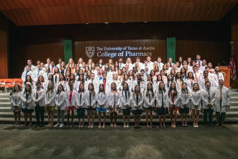 A large group of people in white coats standing in front of the UT Austin College of Pharmacy wordmark.