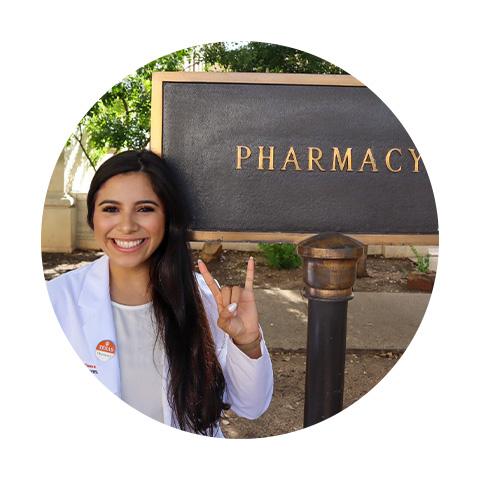 A woman standing in front of the Pharmacy sign and giving the Hook 'em Horns hand gesture.