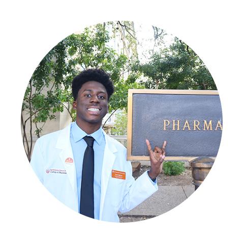 A man wearing a white coat and tie and giving the Hook 'em Horns hand gesture in front of the Pharmacy sign.