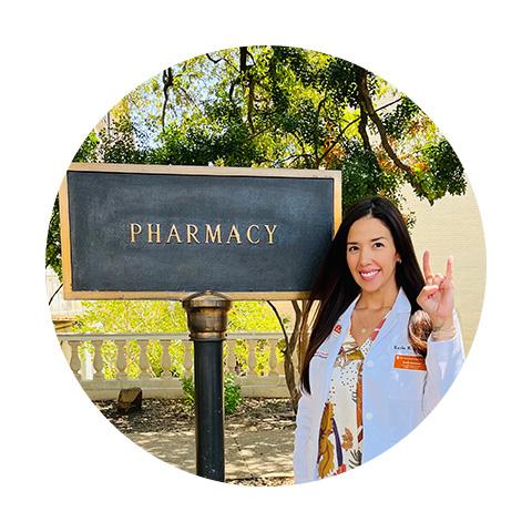 A woman standing in front of the Pharmacy sign and giving the Hook 'em Horns hand gesture.
