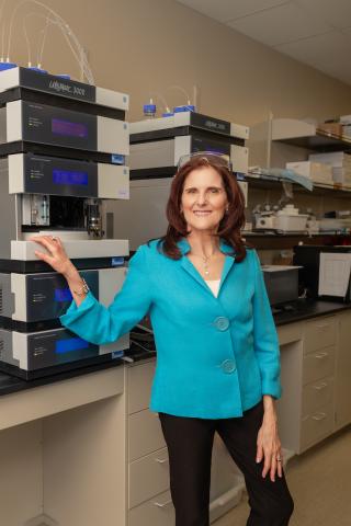 Photo of Dr. Janet Walkow. Walkow is standing in a lab wearing a blue button-up blouse and black pants. She is has shoulder-length brown hair and is smiling to camera.