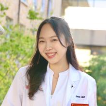A headshot of student pharmacist Doeun Kim. She is wearing a white coat and smiling to camera in a photo taken outside in the courtyard. She has long brown hair.