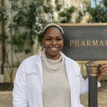 A headshot of Amaka Epoh. Amaka is wearing a white coat and a sparkly beige top. She is standing in front of the Pharmacy building sign and smiling brightly to camera.