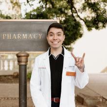A headshot of student pharmacist Adam Campo. Adam is wearing a white coat and black polo shirt, smiling to camera. He is standing by the Pharmacy sign outside of the building on campus and is holding up the "hook 'em horns" UT Austin sign with his hand.