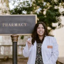 A headshot of student pharmacist Sydney S. Sydney is wearing a white coat and holding her hands up to show the UT Austin "Hook 'Em" sign. She smiles brightly in front of the Pharmacy Building sign on campus.