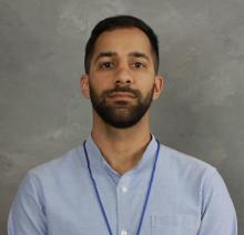 A headshot of Dr. Suheib Omran. He has dark brown hair and a brown beard and is looking directly to camera. Dr. Omran is wearing a light blue shirt and has a blue-corded name tag or lanyard around his neck.