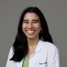 Headshot of student pharmacist Esther. Esther is wearing a white coat and sits smiling brightly at the camera. She has long dark brown hair and is pictured against a grey backdrop.