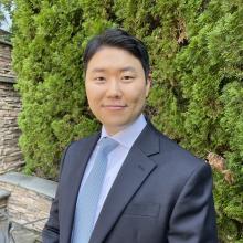 A headshot of Ted Sohn. Ted has black hair and is wearing a grey suit with a blue tie. He is smiling to camera with green shrubbery in the background.