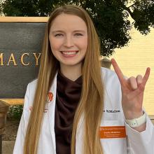 Emilee McMellon smiling in front of the Pharmacy sign and giving Hook 'Em hand sign