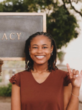 Headshot of Pharmacy student Delandra R. Delandra is wearing a burgundy t-shirt and smiles brightly to camera while holding up a "Hook 'em Horns" hand gesture. She is standing outdoors, in front of the PHARMACY sign on campus.