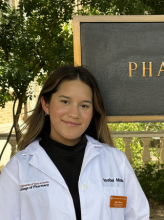 Headshot of pharmacy student Isabel M. Isabel is wearing a black shirt and white lab coat and smiles softly to camera standing in front of the PHARMACY sign on campus.