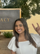 Headshot of pharmacy student Sarah B. Sara is wearing a white, flutter-sleeve dress and is holding up her hand in a "Hook 'em Horns" hand gesture. She smiles brightly to camera, standing in front of the PHARMACY sign on campus.