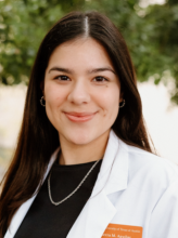 Headshot of Pharmacy student Sierra A. Sierra is wearing a black shirt and chain necklace beneath her white lab coat. She smiles to camera.