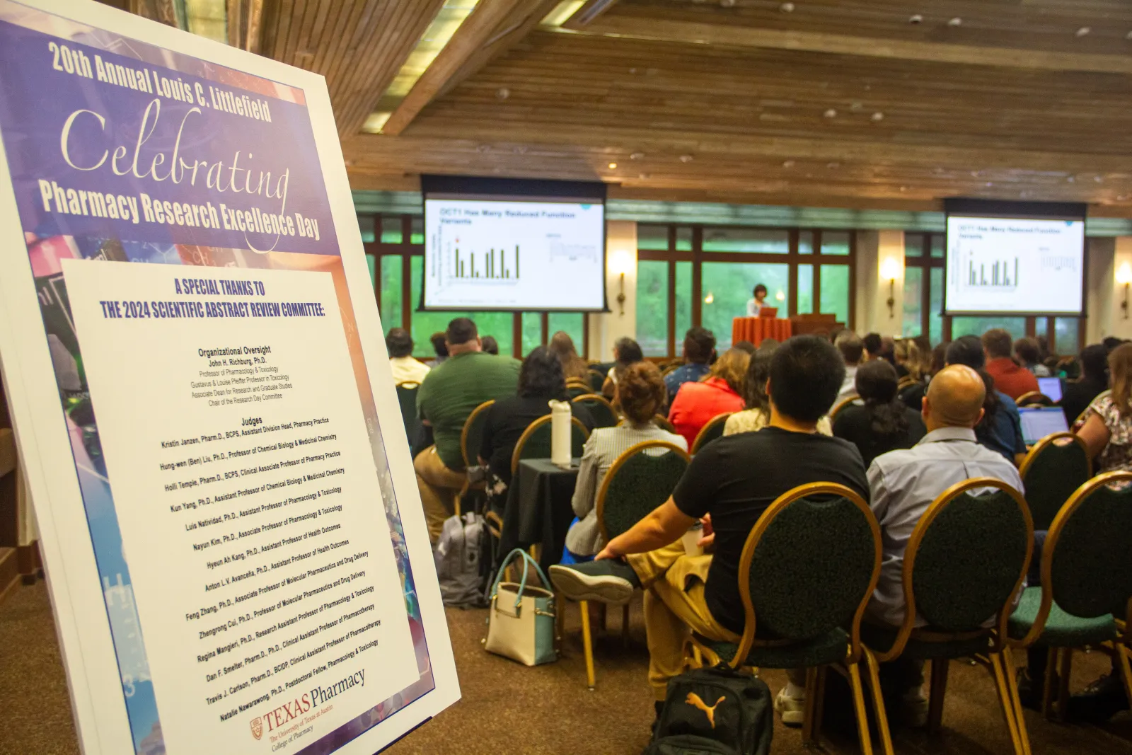 A group of people watching a presentation with a poster on an easel in the foreground.