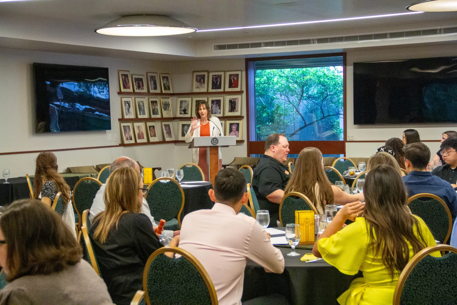 A woman speaking at a podium to a group seated at round tables.