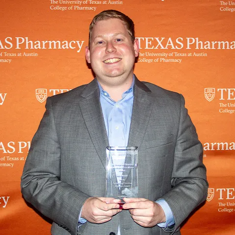 A man wearing a dress shirt and suit coat and smiling while holding an award.