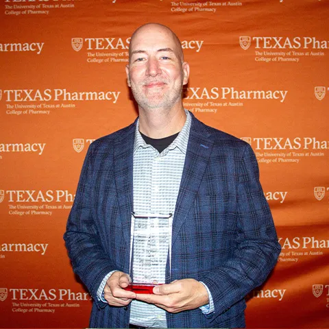 A man wearing a suit jacket and collared shirt and smiling while holding an award.