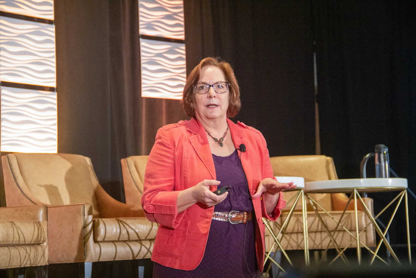 Dr. Sharon Rush stands in front of the stage giving her presentation at PPS. She's wearing a purple dress with a brown belt, a coral-colored blazer and is holding a slide clicker.