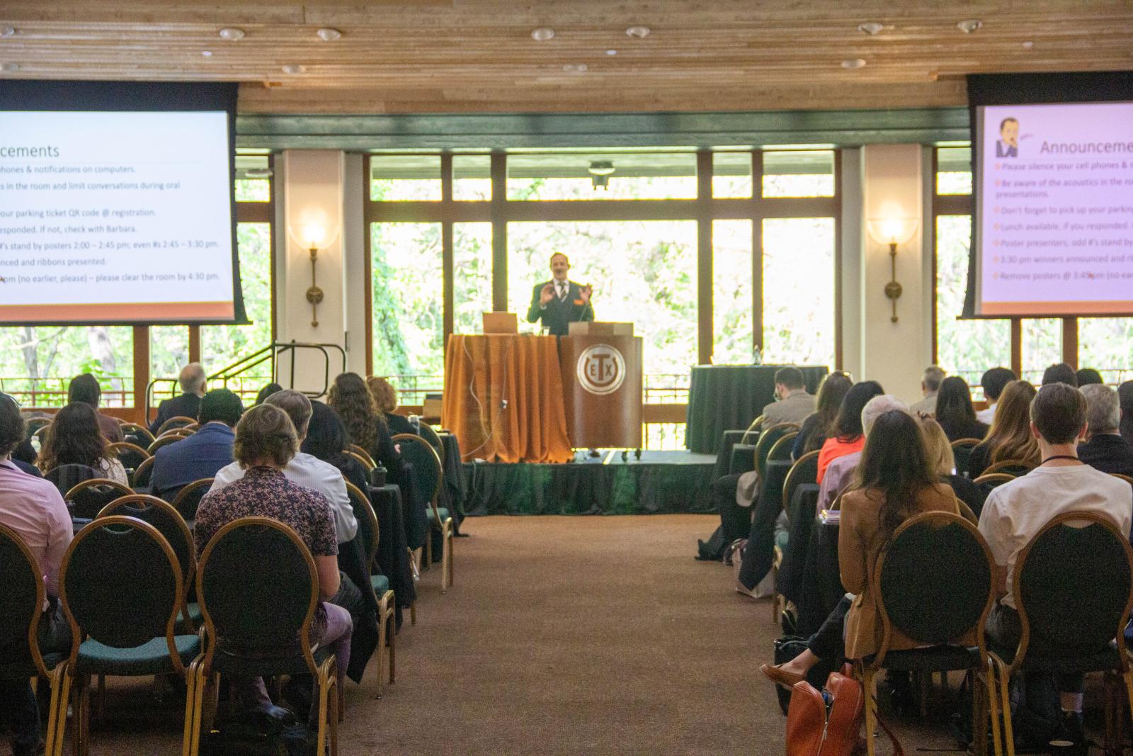 A man presenting at a podium in front of a group of people in a room with large windows.