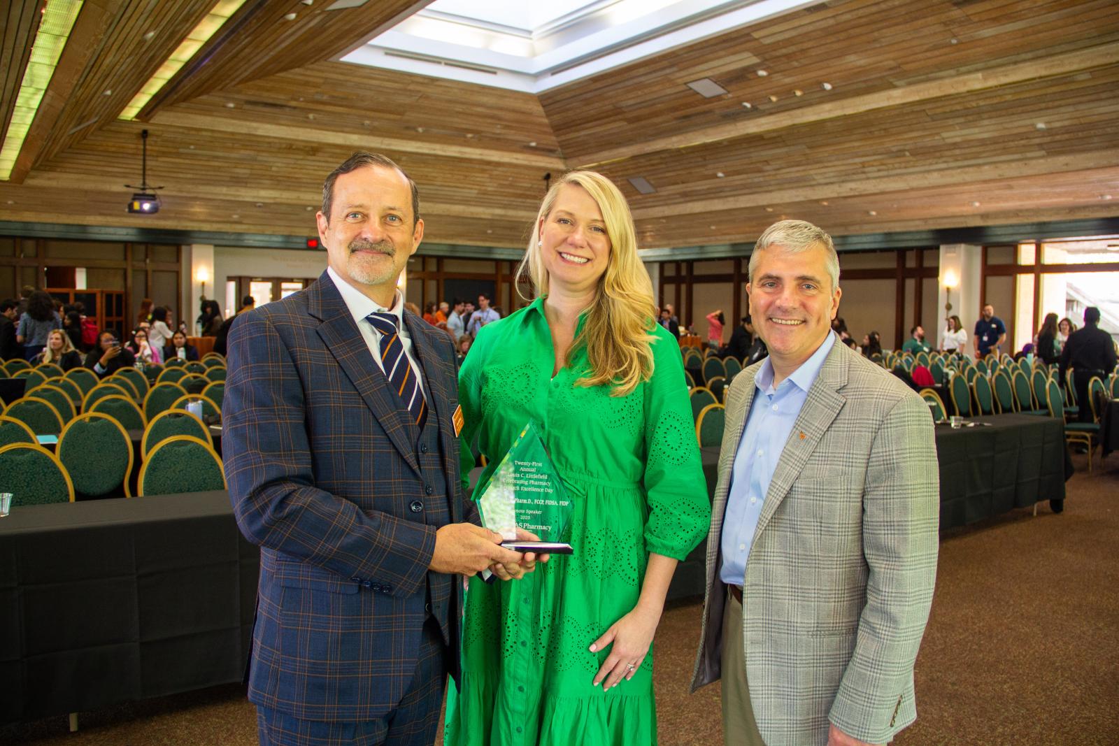 Two men presenting an award to a woman in a large presentation room.