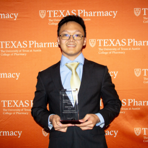 Anthony Wang standing with his award. He is wearing a blue shirt and yellow tie under a black suit coat. He stands in front of an orange, Texas Pharmacy-branded wall.