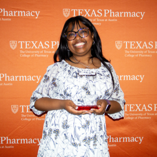 Bri standing with her award. She is wearing a white and black patterned dress and is smiling to camera. Bri stands in front of an orange, Texas Pharmacy-branded wall.