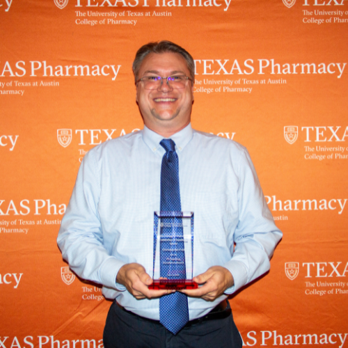 Dr. Frei holding an award. He is wearing a white button-down and blue tie and is standing in front of an orange, Texas Pharmacy-branded wall.