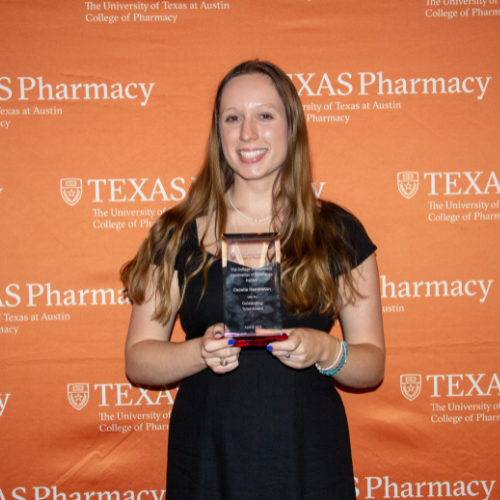 Cecelia standing with her award. She is wearing a black dress and has long blonde-red hair. She stands in front of an orange, Texas Pharmacy-branded wall.
