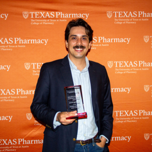 Dan Rivera standing with his award. He is wearing a light blue shirt under a dark suit jacket. He stands in front of an orange, Texas Pharmacy-branded wall.