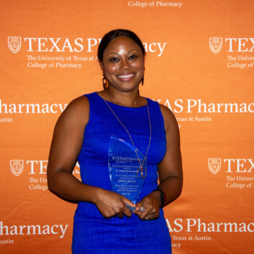 Dr. Lipscomb stands with her award. She is wearing a cobalt blue dress and smiling to camera in front of an orange, Texas Pharmacy-branded wall.