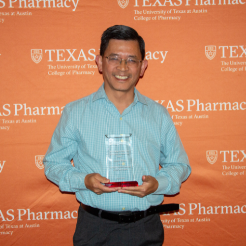 Dr. Pham standing with his award. He is wearing a blue button up shirt and black pants and standing in front of an orange, Texas Pharmacy-branded wall.