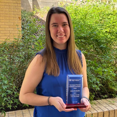 Dr. Summerlin holding her award. She is wearing a royal blue shirt and smiling to camera. Dr. Summerlin is standing outside beside a brick wall and green foliage.