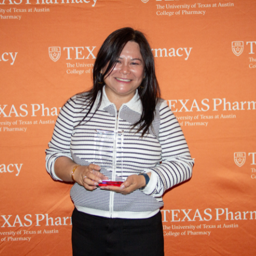 Dr. Dinh standing with her award. She is wearing a striped black and white collared shirt with black pants. She is standing in front of an orange, Texas Pharmacy-branded wall.