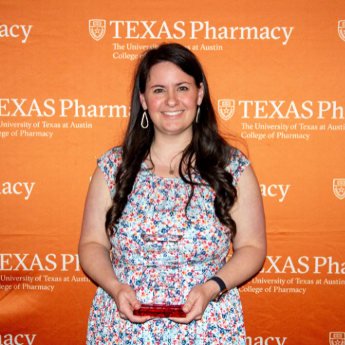 Dr. Kristin Janzen holding an award. She is wearing a floral dress and is standing in front of an orange, Teas Pharmacy-branded wall.